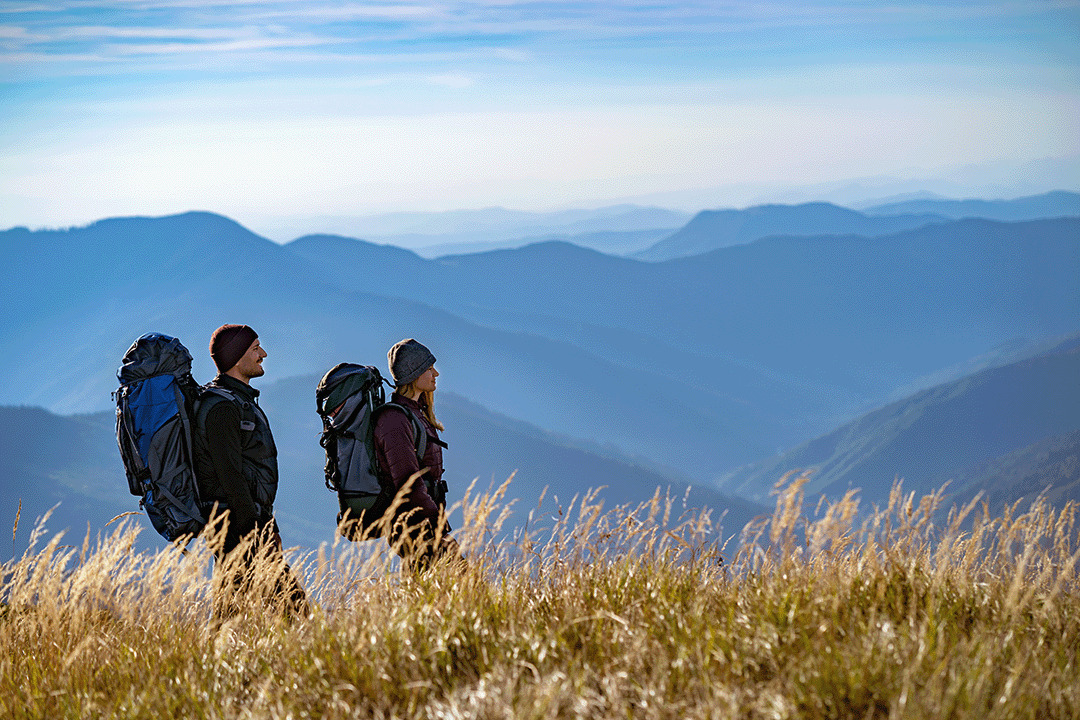 Two hikers with backpacks standing on a mountain top looking at the view.