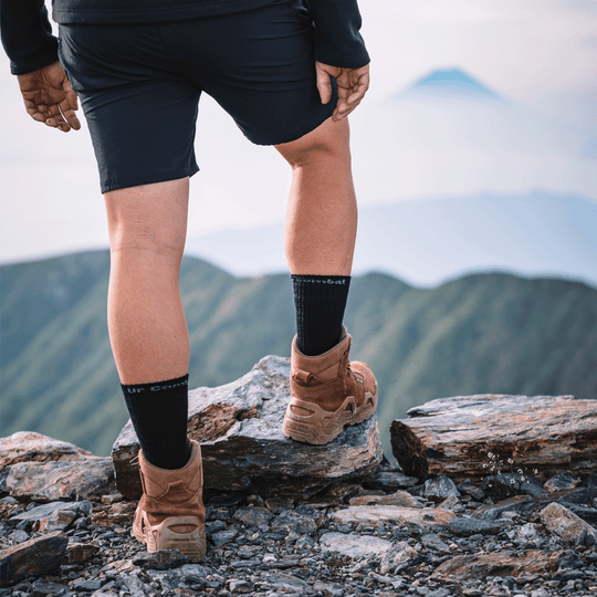 A person hiking on a rocky mountain peak, viewed from behind, wearing black shorts, black combat tactical socks with white lettering, and tan lace-up boots. The background shows blurred green mountain ridges and a hazy sky.