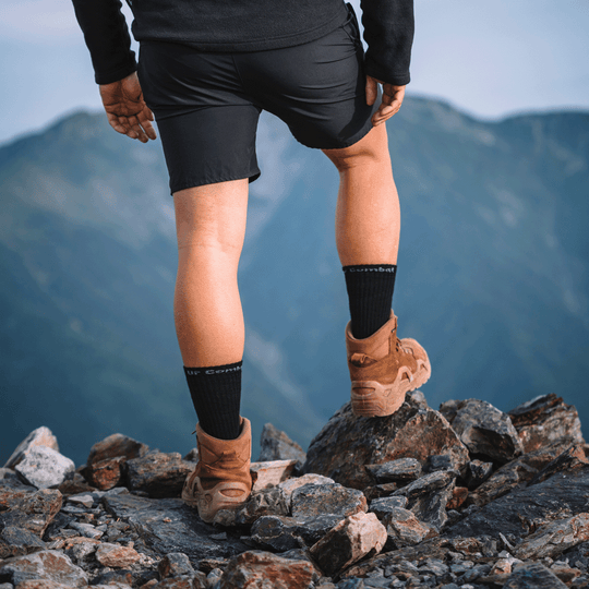 A person hiking on rocky mountains wearing black athletic shorts, black combat tactical socks with white branding, and brown hiking boots, shown from behind.