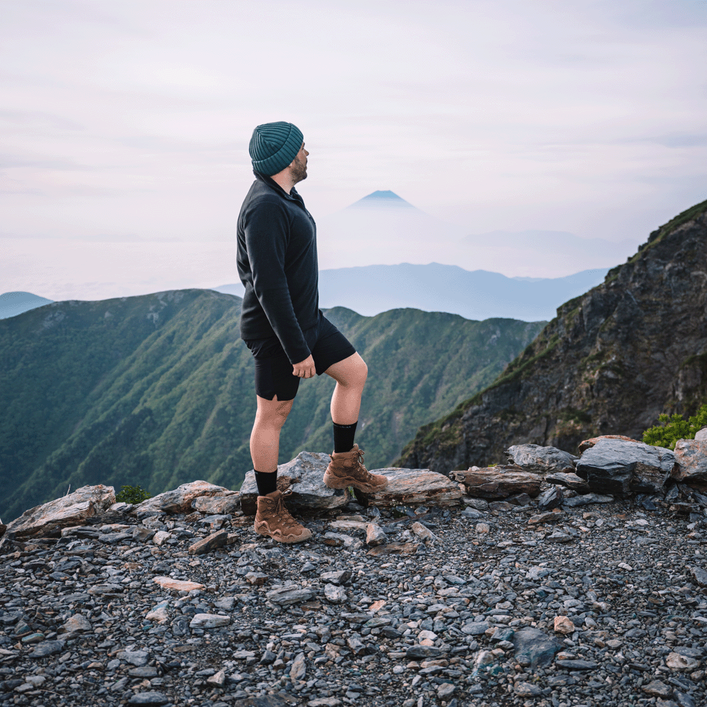 A man stands on a rocky mountain ridge wearing a dark knit beanie, black long-sleeve top, black shorts, and brown hiking boots with black combat tactical socks, looking out over layered green mountain ranges under a pale sky.