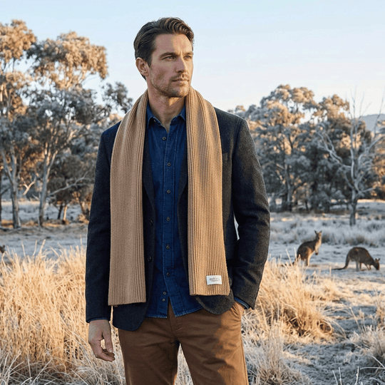 Man standing in a field with kangaroos, wearing a dark jacket, blue shirt, and brown scarf.