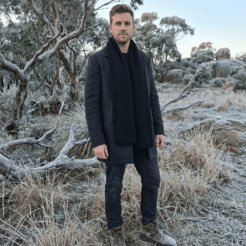 Man standing in a frosty, snowy landscape wearing a dark coat and scarf.