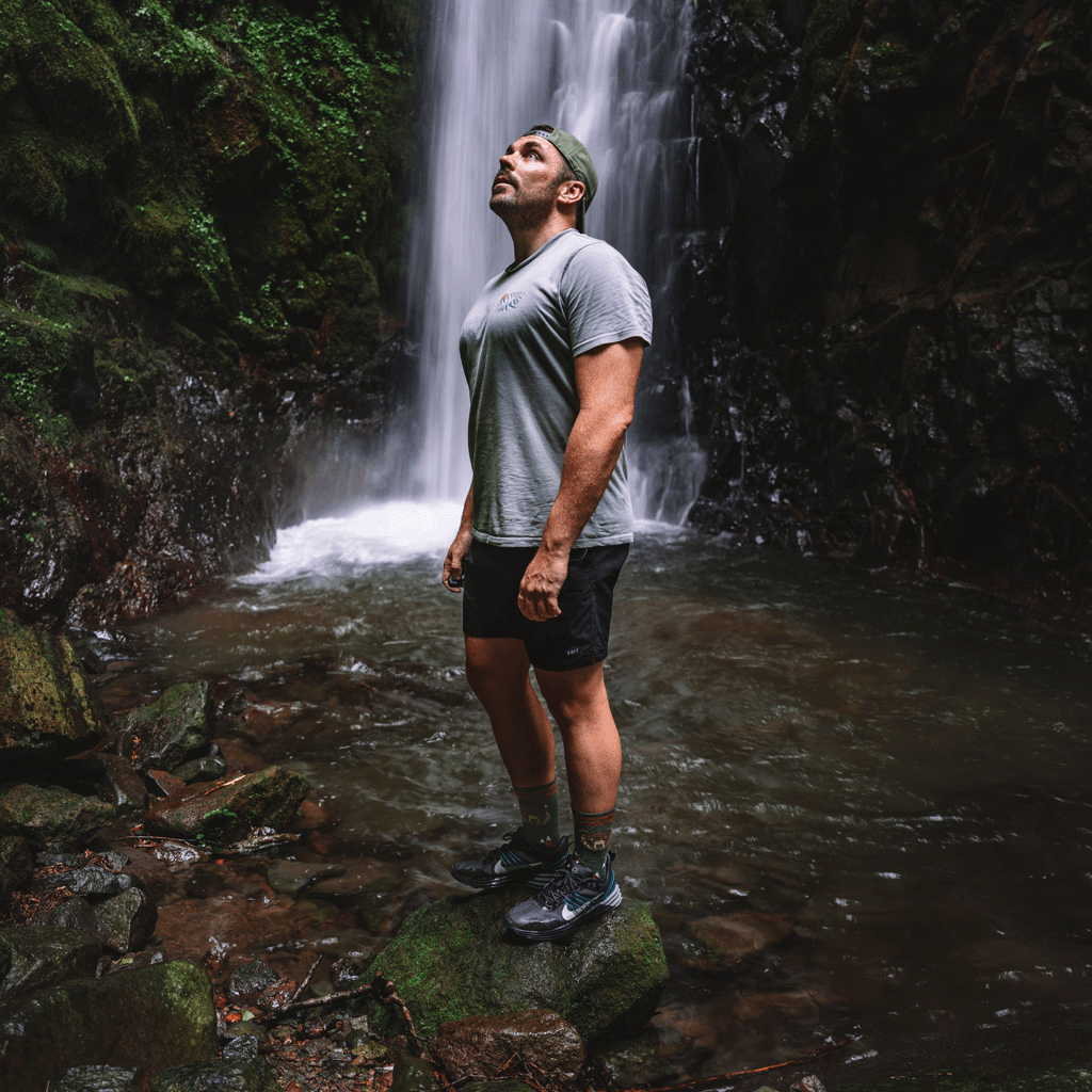 A man stands in a rocky stream in front of a waterfall, wearing a gray t-shirt, black athletic shorts, green lifestyle socks, and black running shoes.