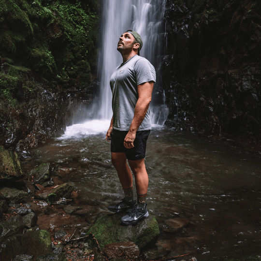 A man stands in a rocky stream in front of a waterfall, wearing a gray t-shirt, black athletic shorts, green lifestyle socks, and black running shoes.