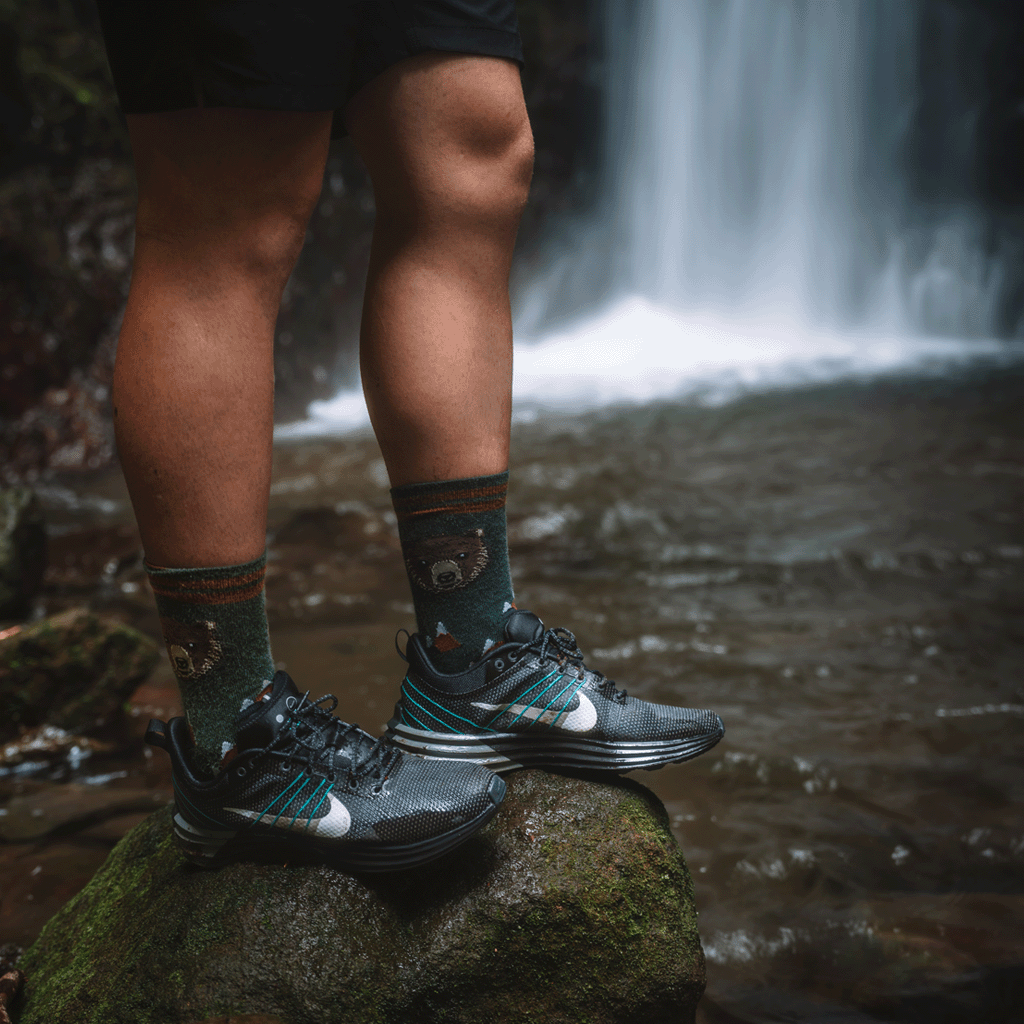 A person standing on a mossy rock by a waterfall, wearing black hiking shoes with teal and white accents, dark patterned lifestyle crew socks, and black shorts.