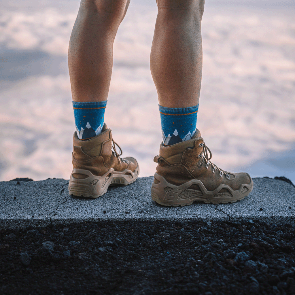 A person standing outdoors on a concrete ledge wearing tan hiking boots and bright blue crew socks with a white mountain pattern.