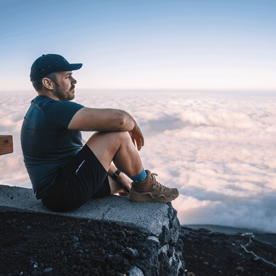 A man sits on a stone ledge overlooking a sea of clouds at  sunset, wearing a dark blue t-shirt, black shorts, a black cap, life style socks and brown hiking shoes.