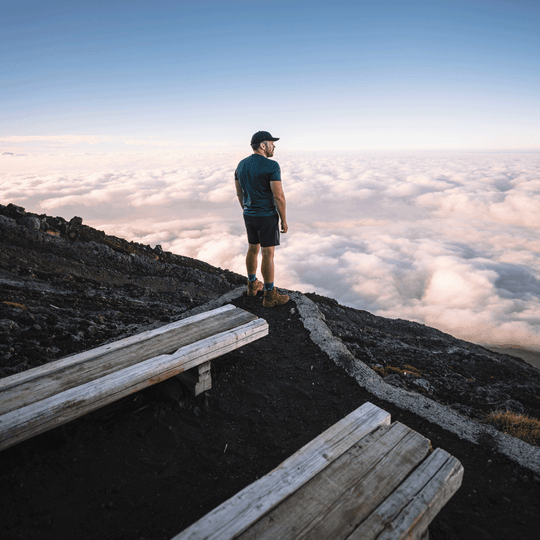 A man stands on a mountain ridge above a sea of clouds, wearing a dark blue t-shirt, black shorts, boots, lifestyle socks and a black cap, with wooden benches in the foreground.