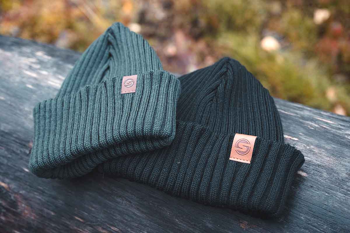One green and one black knit beanies with brown labels on a wooden surface with a blurred natural background