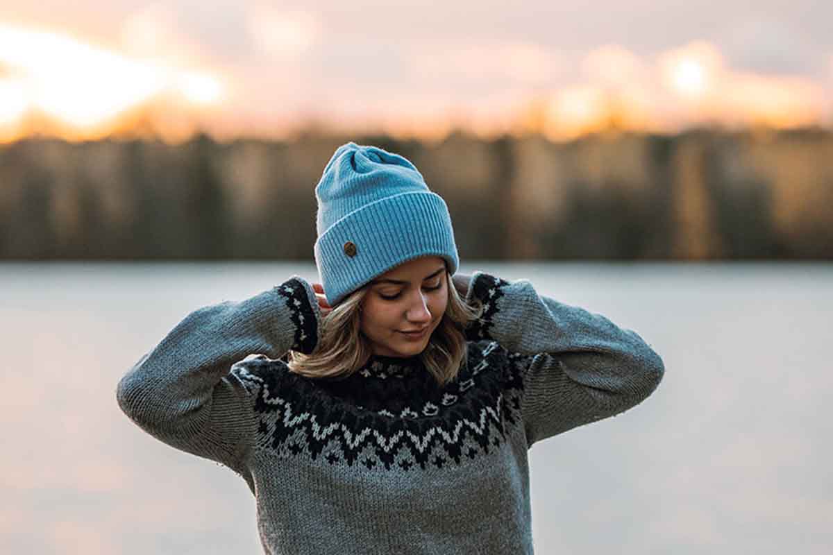 Woman wearing a blue knit beanie and gray sweater with a patterned collar by a lake at sunset.