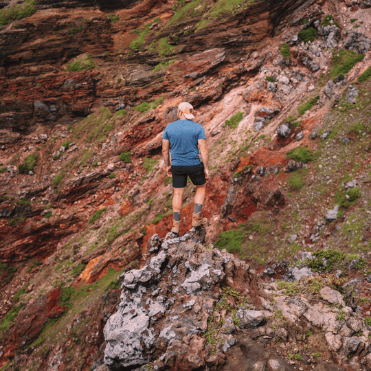 A man wearing a blue t-shirt, black shorts, hiking boots and grey Posio trekking socks stands on a rocky outcrop, looking out over a steep, rugged mountain slope with reddish-brown cliffs and patches of green vegetation.