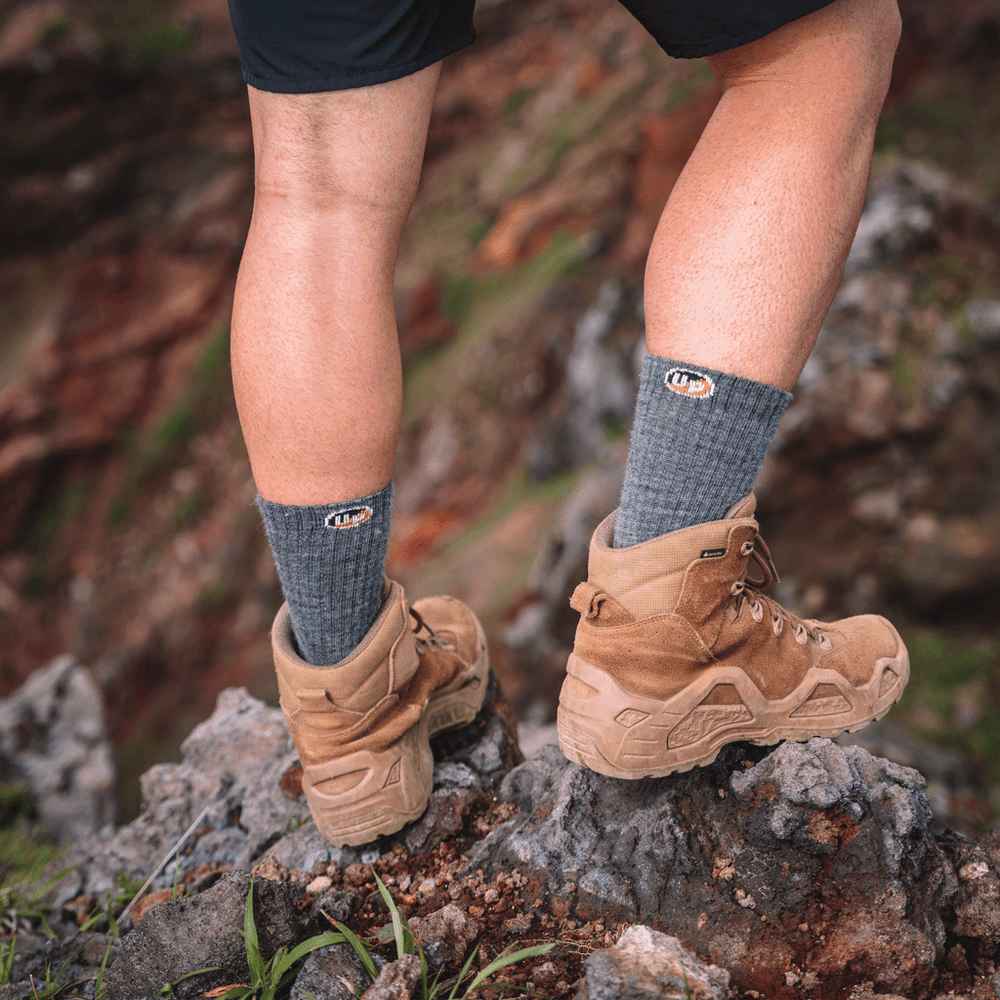 A close-up outdoor shot of a person's legs wearing brown tactical boots and gray Posio Trekking socks with a visible UphillSport logo, standing on a rocky cliffside.