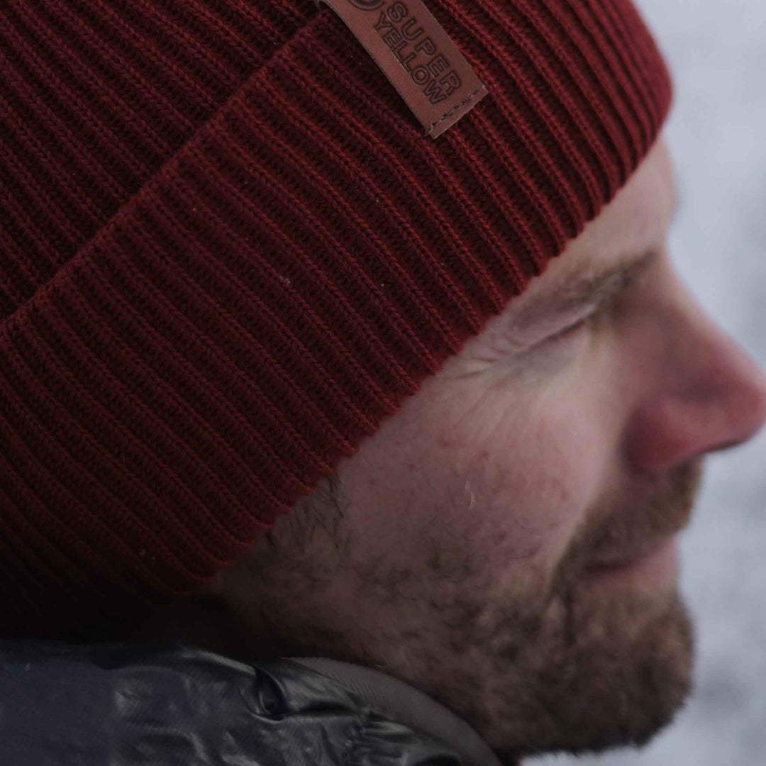 A red-coloured merino wool beanie with a thick knit pattern and a small brown brand tag attached to the brim.